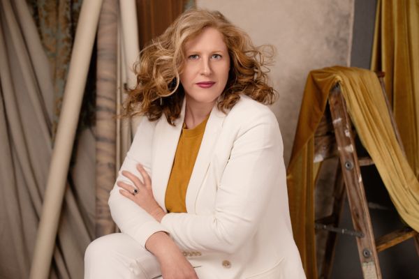 Jennifer in white suit and gold top sits in front of a studio backdrop draped in different fabrics.