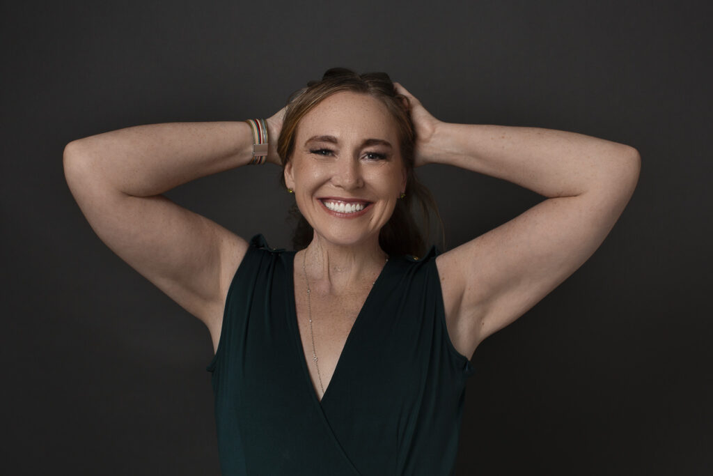 An editorial portrait of a woman in a green jumpsuit standing confidently against a dark background, arms raised behind her head.