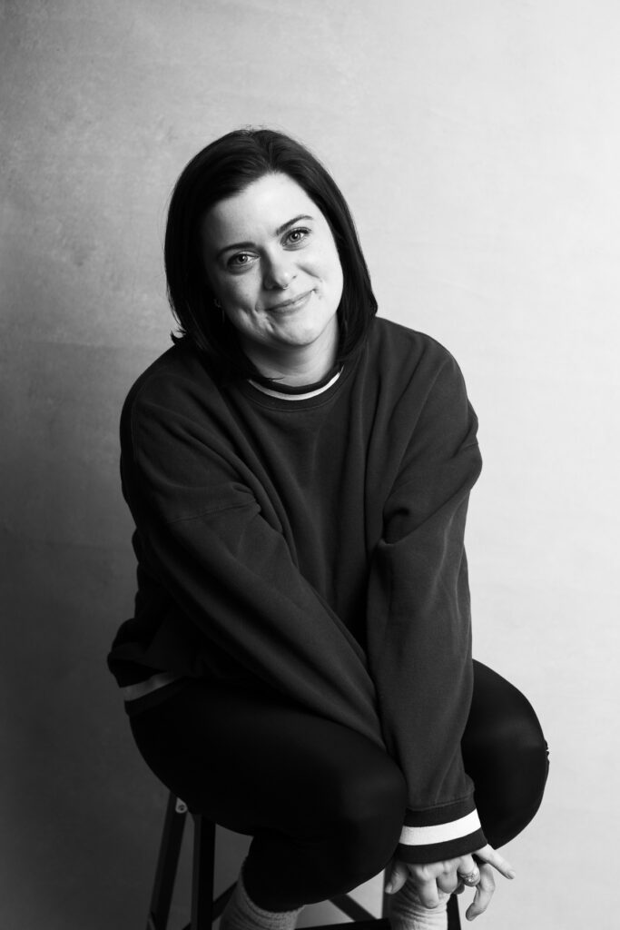 Woman in sweatshirt is seated on a stool and clasps hands between knees. She smiles at the camera in a studio portrait at Redhead Creative Media in Chapel Hill, NC.