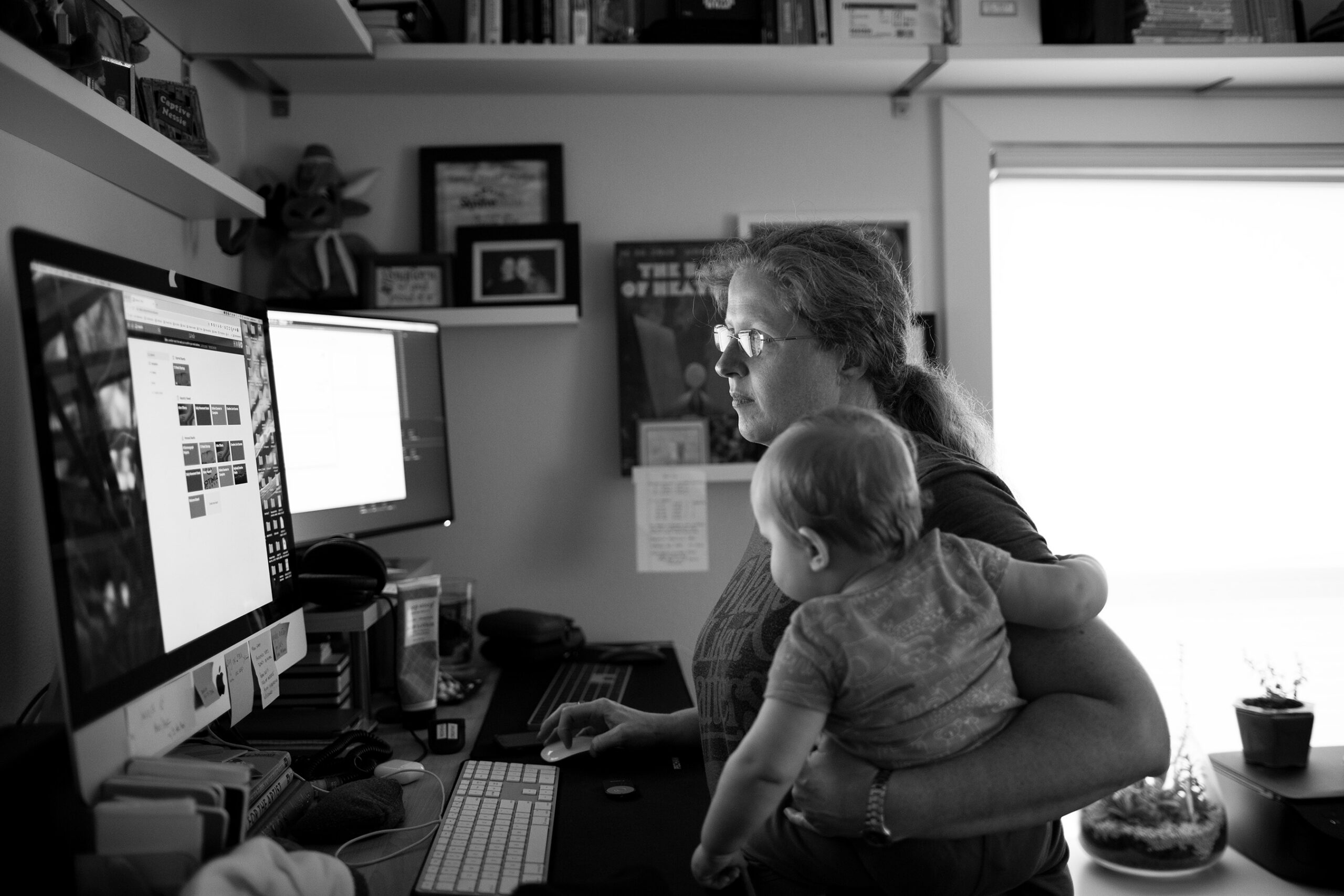 A black and white documentary photograph of Jennifer Horst standing at her home office desk, holding a baby on her hip with one hand while working at dual computer monitors with the other, shelves of books and framed art visible behind her, natural window light falling across the scene.