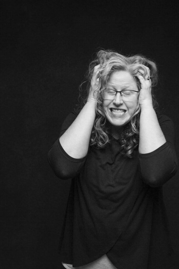 A black and white portrait of Jennifer Horst against a dark background, both hands gripping her hair, eyes closed and expression raw. This is an honest, unguarded image of overwhelm captured as part of her Postpartum Project self-portrait series.